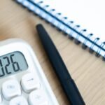 Calculadora exibindo 2026 sobre mesa de madeira, ao lado de um caderno espiral e uma caneta — imagem associada a planejamento financeiro, atualização de valores e cálculos de RPVs e precatórios.