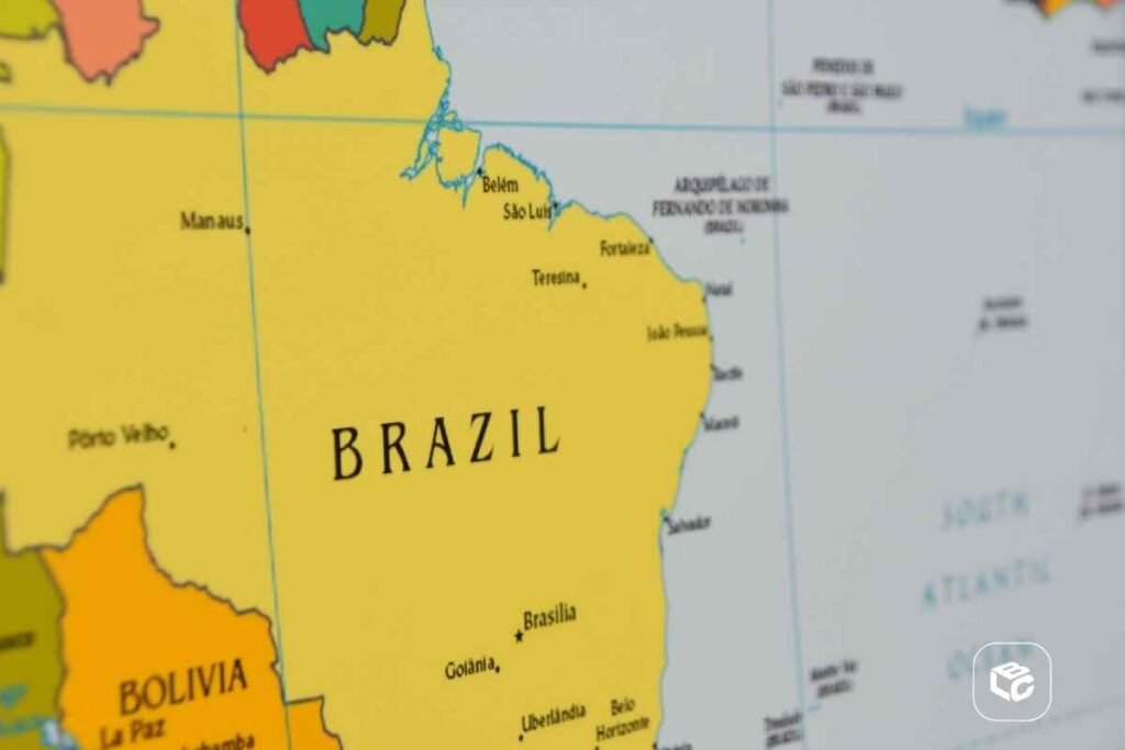 Mapa do Brasil em destaque, exibindo parte da América do Sul e o Oceano Atlântico. O território brasileiro aparece em amarelo, com o nome “BRAZIL” ao centro. Diversas cidades estão identificadas no mapa, incluindo Brasília, Goiânia, Manaus, Belém, São Luís, Teresina, Fortaleza, João Pessoa, Natal, Recife, Maceió, Aracaju, Salvador, Belo Horizonte e Curitiba. Também é possível ver a Bolívia destacada em laranja à esquerda, com a cidade de La Paz indicada. O mapa possui linhas de grade geográfica em azul-claro e um logotipo branco no canto inferior direito da imagem.