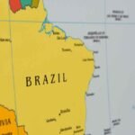 Mapa do Brasil em destaque, exibindo parte da América do Sul e o Oceano Atlântico. O território brasileiro aparece em amarelo, com o nome “BRAZIL” ao centro. Diversas cidades estão identificadas no mapa, incluindo Brasília, Goiânia, Manaus, Belém, São Luís, Teresina, Fortaleza, João Pessoa, Natal, Recife, Maceió, Aracaju, Salvador, Belo Horizonte e Curitiba. Também é possível ver a Bolívia destacada em laranja à esquerda, com a cidade de La Paz indicada. O mapa possui linhas de grade geográfica em azul-claro e um logotipo branco no canto inferior direito da imagem.